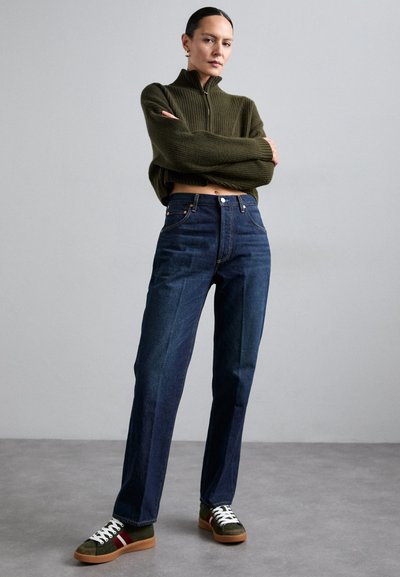 Dunkelblaue hochtaillierte Jeans, kombiniert mit einem olivgrünen strukturierten Pullover und grünen Turnschuhen mit roten und weißen Akzenten.
