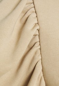 Beige textil med ribbad textur, prydd med hopskrumpna volanger vid sömmen, vilket skapar dimension och visuell intresse i tyget.