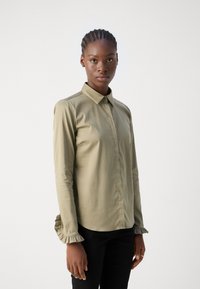 Camisa de manga longa verde oliva com colarinho abotoado, punhos franzidos, textura suave e design ajustado. Usada com calças pretas.