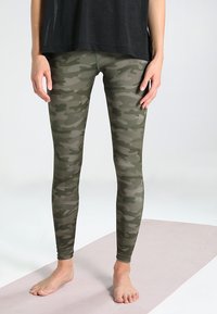 Leggings de camuflaje en tonos verdes, hechos de tela elástica, que presentan un diseño ajustado y una textura suave, combinados con una blusa negra suelta.
