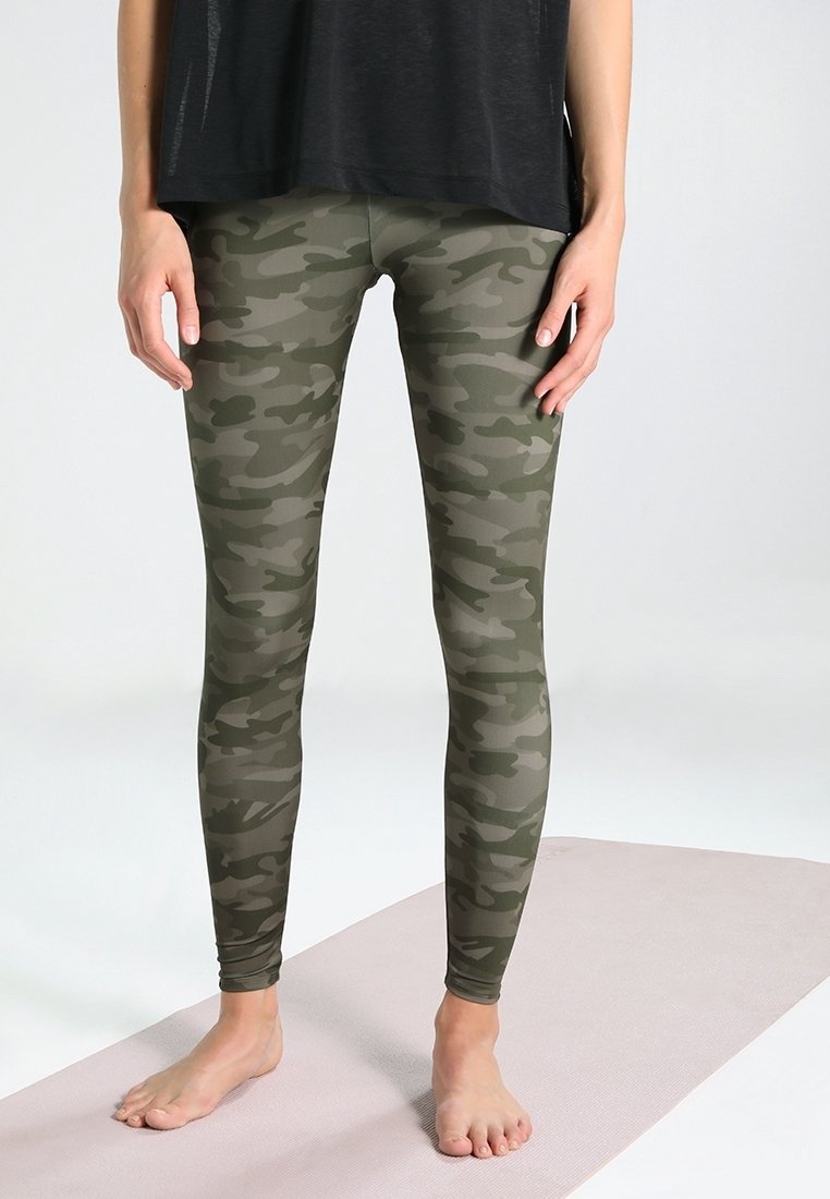 Leggings de camuflaje en tonos verdes, hechos de tela elástica, que presentan un diseño ajustado y una textura suave, combinados con una blusa negra suelta.