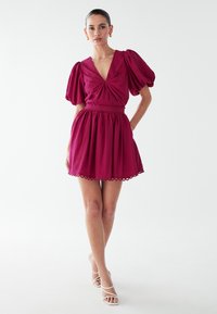 Přiléhavé vínové šaty s nabíranými rukávy, výstřihem do V, seřazeným trupem a rozšířenou sukní. Mají osmihranný lem a úzký pas.