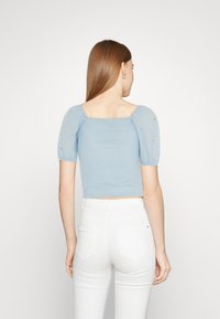 Ανοιχτό μπλε cropped τοπ με κοντά φουσκωτά μανίκια από κεντημένο ημιδιάφανο ύφασμα; εφαρμοστό ριγωτό σώμα. Μοντέλο που φοράει λευκά παντελόνια.