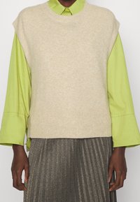 Person, die einen beigen gestrickten ärmellosen Pulli über einem hellgrünen Langarmshirt und einem plissierten dunklen metallischen Rock trägt, steht vor einem weißen Hintergrund.