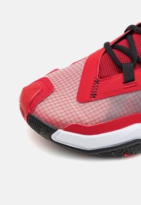 Chaussure de sport rouge et noire avec motif en mesh et grille sur l’orteil, lacets noirs, semelle intermédiaire blanche et semelle extérieure noire texturée.