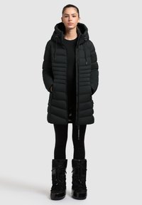 Čierna prešívaná bunda s horizontálnym vzorom, kapucňou a zipsom. Kombinovaná s čiernymi legínami a lesklými čiernymi čižmami.