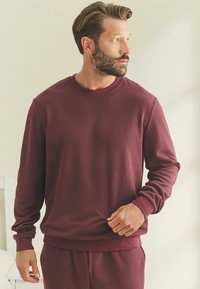 Bordeaux színű, strukturált pulóver hosszú ujjakkal, kerek nyakkivágással és bordás mandzsettákkal. Az anyag puhanak tűnik, finom mintával és laza szabással rendelkezik.