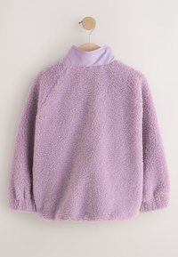 Lavendel lilla fleecetrøje med høj krave hængende på en træbøjle mod en plain væg, der viser bagsiden.