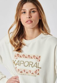 Crème sweater met metallic gouden en glinsterende bloemenprint met de tekst "KAPORAL" en "TIJD OM DE WINTER TE VERGETEN." Gezellige pasvorm.