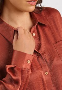 Woman's hand adjusting the collar of a shiny rust-colored button-up shirt with beige buttons and a pocket on the chest.