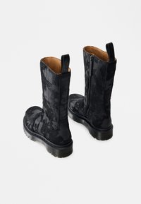 Melni puszābaki līdz vidējai teļiem ar teksturētu ziedu rakstu, sānu rāvējslēdzēju, ievelkamu cilpu un biezām caurspīdīgām zolēm uz balta fona.