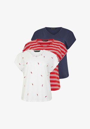 Kobieta o długich blond włosach, ubrana w białą koszulkę z dekoltem w serek ozdobioną nadrukami czerwonych papryczek chili oraz niebieskie dżinsy, stojąca na jasnym, jednolitym tle.