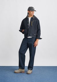 Σκούρο μπλε τζάκετ με κίτρινη λεπτομέρεια στο φερμουάρ, λευκό μπλουζάκι, σκούρες μπλε παντελόνες, και μπεζ μπότες. Ένα γκρι καπέλο ολοκληρώνει το σύνολο.