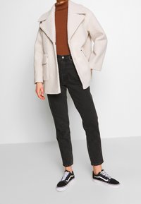 Abrigo de mezcla de lana texturizada en color beige con solapas sobredimensionadas y bolsillos laterales, combinado con jeans oscuros de tiro alto y zapatillas negras.