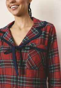 Czerwony flanelowy top piżamy w kratkę z granatowym falbanowym kołnierzykiem, wiązaniem oraz kieszenią. Wykonany z miękkiego materiału, z przecinającymi się zielonymi i białymi liniami.