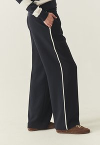 Pantalon large bleu marine en tissu doux, avec une bande latérale blanche contrastante et des poches sur les côtés. Associé à des chaussures marron.