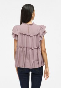 Blusa viola con maniche corte arricciate e design a strati. Presenta una chiusura a forma di chiave sulla schiena e un tessuto morbido e leggero.