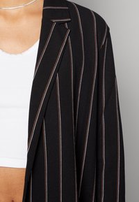 Svart kavaj med vertikala ränder i orange och vitt, med en skräddarsydd design, mjuk struktur och snibbkrage, bärs över en vit topp.