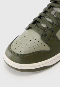 Grøn lædersneaker med perforeret tå, olivengrønne accenter, flade snørebånd og hvid mellemsål. Tekstureret overflade og syede detaljer er synlige.