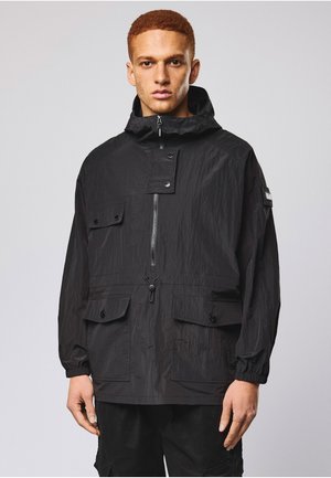 Ung mand iført en sort hættetrøje med lynlås foran, klaplommer og elastiske manchetter, stående foran en simpel lysegrå baggrund.