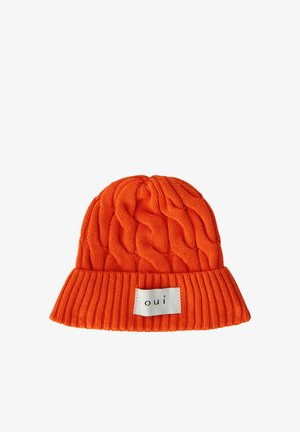 Hellorange gestrickte Mütze mit dicker Zopfmusterung und umgeschlagenem geripptem Rand mit einem weißen Aufnäher mit dem Schriftzug "oui" in schwarzem Text.