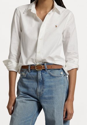 Frau trägt ein weißes Hemd mit hochgekrempelten Ärmeln, das in eine hoch taillierte hellblaue Jeans gesteckt ist, dazu einen braunen Gürtel als Accessoire.