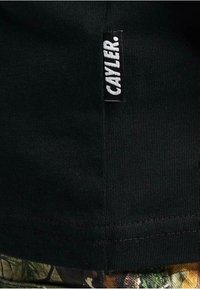 Sort bomulds T-shirt med en struktureret overflade og et hvidt vævet mærke med teksten "CAYLER" på sidesømmen.
