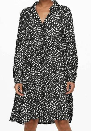 Zwarte jurk met lange mouwen met witte onregelmatige polkadots, V-hals, losse pasvorm en gelaagde rok, gedragen door een staande persoon.