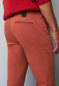 Chino-broeken in terracotta, met achterzakken, een gladde textuur en een zwart merklabel op de tailleband.