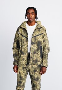 Camouflage ydertøj i nuancer af grøn og brun, med hætte, lynlås lommer og et afslappet fit design.