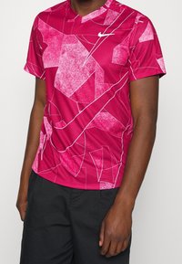 Czerwona sportowa koszulka z geometrycznym wzorem oraz nieregularnymi białymi akcentami, krótkimi rękawami i okrągłym dekoltem. Logo Nike na piersi.