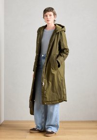 Olijfgroen parka met een ritssluiting aan de voorkant, grote zakken en een verstelbare capuchon; gecombineerd met lichtblauwe wijde jeans en bruine sneakers.