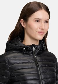 Sort pufferjakke med quiltet design, hætte og frontlynlås. Har justerbare snore til tilpasning og en glat, skinnende overflade.