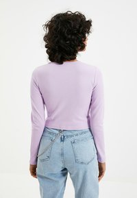 Lila pitkähihaine paita, jossa on pyöreä kaula-aukko, yhdistettynä vaaleansinisiin korkeavyötäröisiin farkkuihin, joissa on koristeellinen hopeaketju.