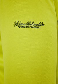 Camicetta gialla a maniche corte realizzata in tessuto testurizzato, con testo ricamato blu: “Schmuddelwedda WENN DAT PLADDERT” vicino alla spalla.