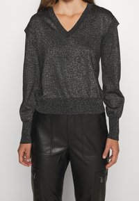 Schwarzer metallischer V-Ausschnitt-Pullover mit langen Ärmeln und geripptem Saum, der einen subtilen Glanz und strukturierte Schulterdetails aufweist, kombiniert mit schwarzen Lederhosen.