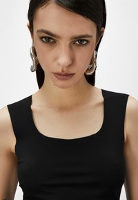 Top nero senza maniche con scollatura quadrata, realizzato in tessuto liscio. La modella indossa grandi orecchini metallici lucidi. Pelle chiara, trucco neutro.