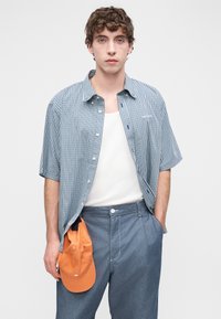 Πουκάμισο με κοντά μανίκια και κουμπιά σε μπλε και λευκό καρό μοτίβο, φορεμένο πάνω από λευκή αμάνικη μπλούζα, συνδυασμένο με τζιν παντελόνια και πορτοκαλί καπέλο.