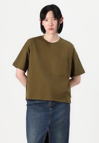 Olivegrünes Baumwoll-T-Shirt mit Rundhalsausschnitt, kurzen Ärmeln und einer Vordertasche. Getragen mit einem hoch taillierten Jeansrock.