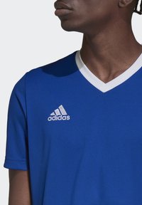 Blå idrettsjersey med V-hals og hvit krage. Har et sølvfarget Adidas-logo på venstre bryst og korte ermer. Glatt tekstur.