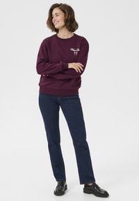 Sweatshirt bourgogne avec col rond et broderie blanche "Wine Club", associé à un jean bleu foncé et des mocassins noirs, se tenant sur un fond uni.