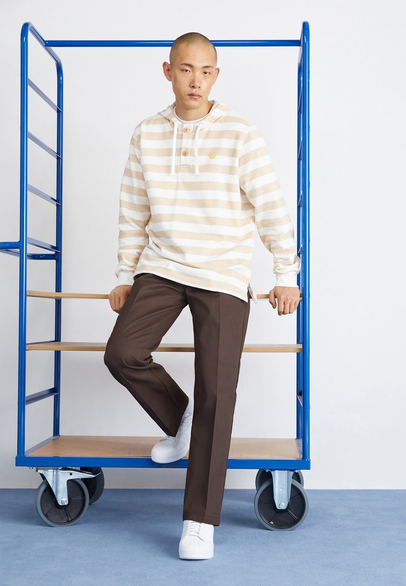 Sudadera a rayas beige y blanca con un bolsillo frontal y botones, combinada con pantalones marrones y zapatillas blancas. El fondo presenta un carrito azul.