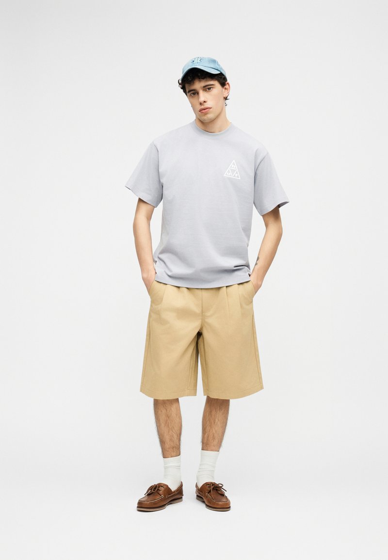 Νεαρός άνδρας που φοράει ανοιχτό γκρι μπλουζάκι, μπεζ σορτς μέχρι τα γόνατα, λευκές κάλτσες, καφέ δερμάτινα παπούτσια και ανοιχτό μπλε καπέλο, στέκεται με τα χέρια στις τσέπες.