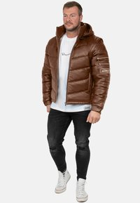 Giacca imbottita marrone con cappuccio, chiusura a zip e molteplici tasche. Indossata sopra una camicia bianca e abbinata a jeans neri e sneakers.