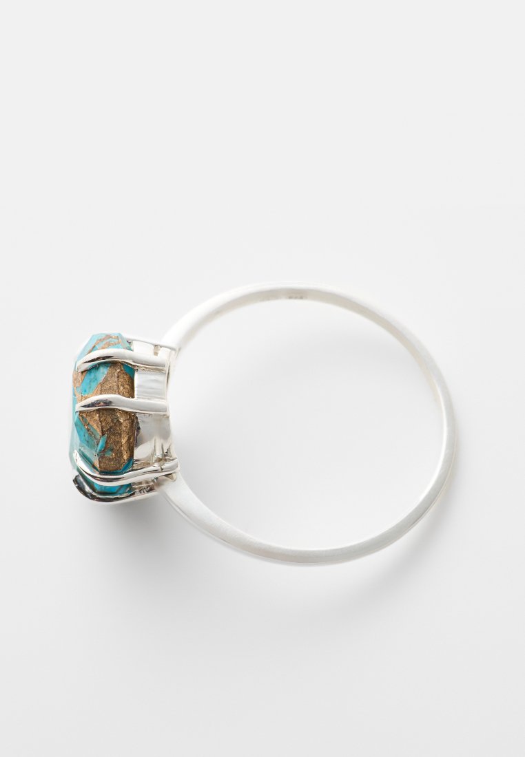 Anillo de plata con una gran piedra irregular de turquesa engastada en un soporte de cuatro garras sobre una banda lisa, contra un fondo blanco.