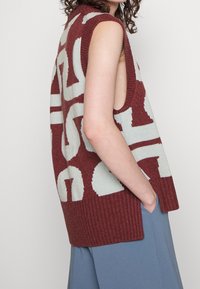 Bezrękawnik dzianinowy w kolorze maroon z dużymi białymi wzorami geometrycznymi. Bocznie rozcięcia zwiększają cyrkulację powietrza i komfort noszenia.