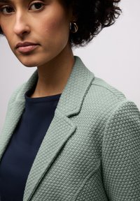 Blazer vert menthe texturé avec un col cranté, associé à un haut bleu marine foncé, présentant une coupe structurée et des détails de tissu complexes.