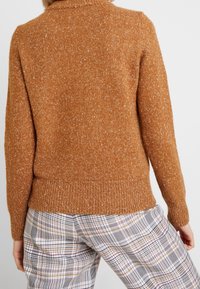 Brauner, strukturierter Pullover mit geripptem Saum und Kragen, getragen über karierter Hose, die verschiedene Farben im Design aufweist.