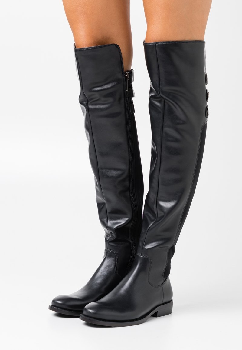 Bottes en cuir noires montant jusqu'au genou avec talons bas et fermeture éclair latérale portées sur jambes nues sur fond blanc.