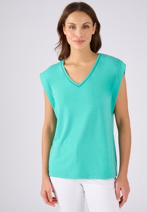 Pull turquoise à col en V avec des manches courtes, texture lisse et coupe décontractée, associé à un pantalon blanc.